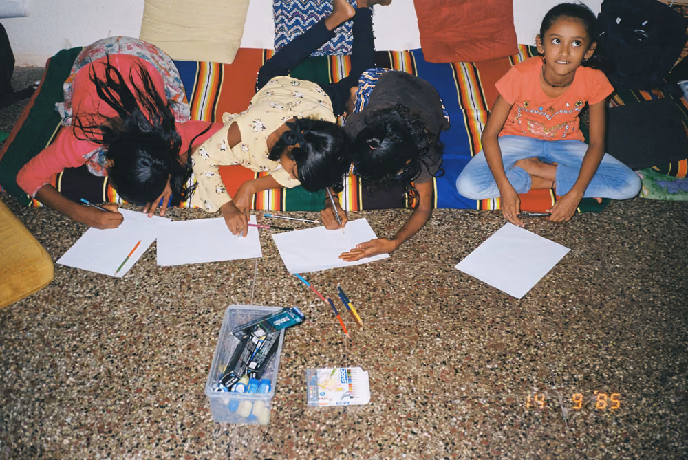 Children lying on the floor drawing on sheets of paper, a box of markers open between them.