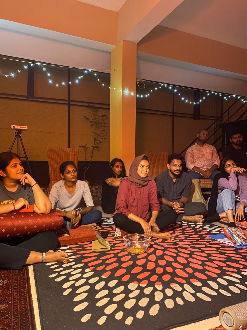 People sitting cross-legged on a patterned rug under a string of fairy lights, listening to someone off camera.