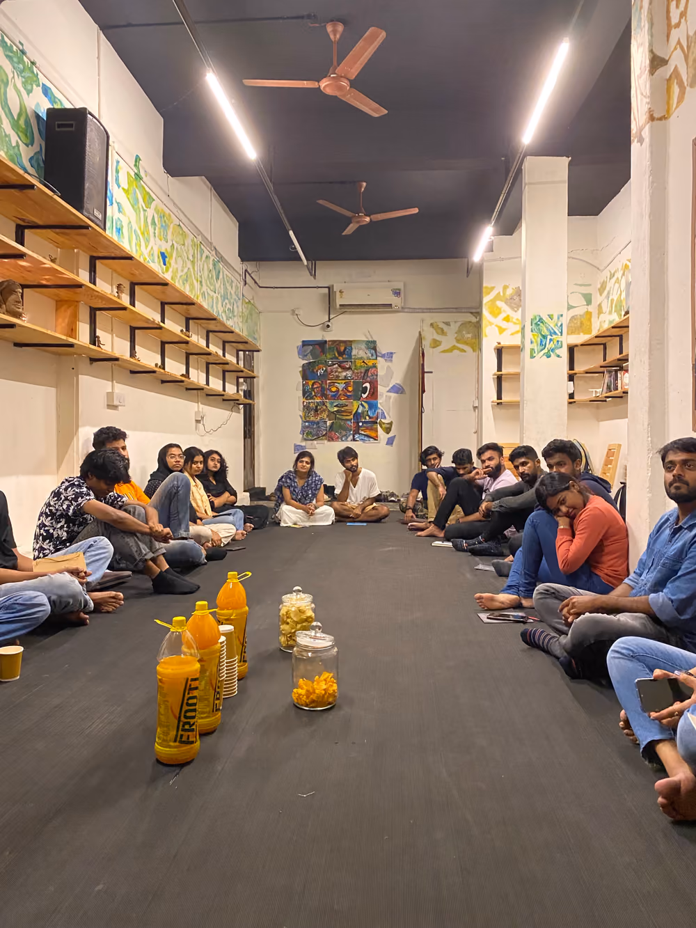 Two rows of people sitting on the floor of a long studio, facing each other across a line of bottles and jars of snacks.
