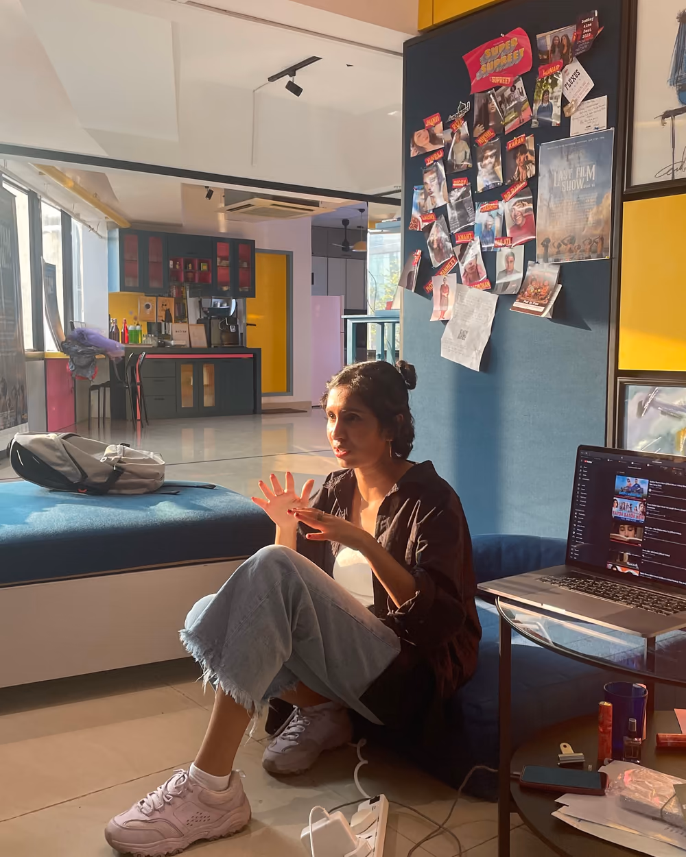 Raiza sitting on the floor beside a laptop, talking with her hands, a wall of pinned-up film posters behind her.
